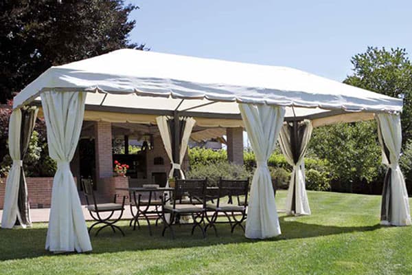 Complementi arredo Didò Floricoltura targati Greenwood, gazebo con telaio in alluminio della serie Nebraska con teli in stoffa bianchi, situato in un giardino con sotto un tavolo con delle sedie in ferro battuto.